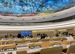 [ai] Interior view of a large conference room with rows of tables and nameplates for various countries. A ceiling mural displays vibrant colors. Attendees are seated, engaged in discussions, while a screen displays information.